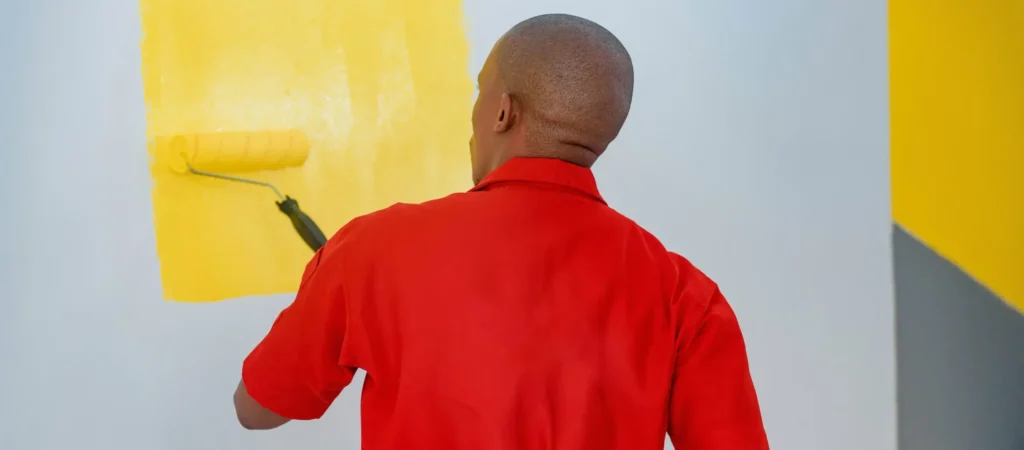 A professional painter in a red jumpsuit applies a vibrant yellow coat to an exterior wall, demonstrating why late spring and early fall are the best time of year to paint your home in Fairfield, CT.