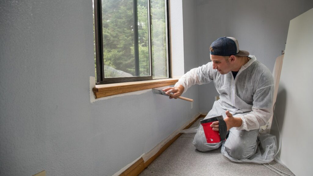 A man in a painter's suit uses a brush to paint a wall section