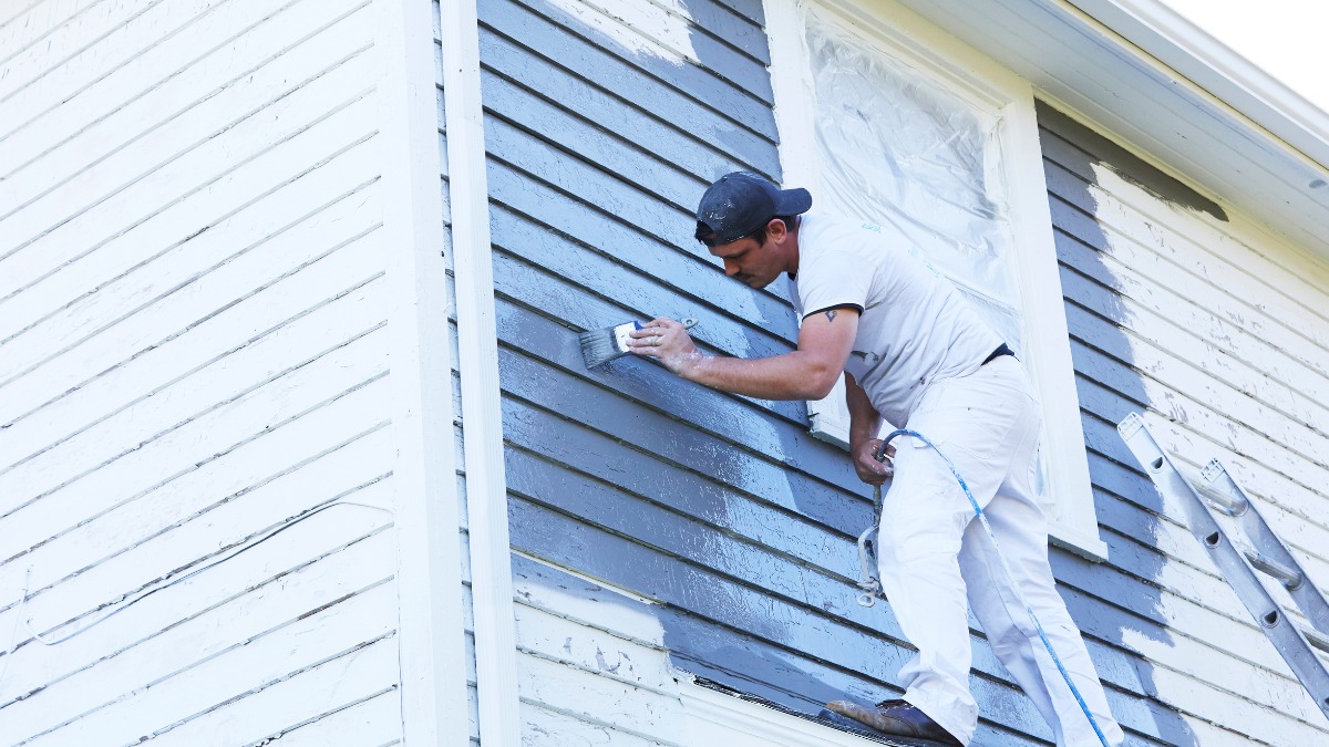 Painter brushing grey paint onto house siding